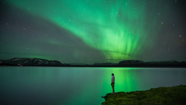 how are the northern lights formed? Man stands at the edge of a lake watching northern lights